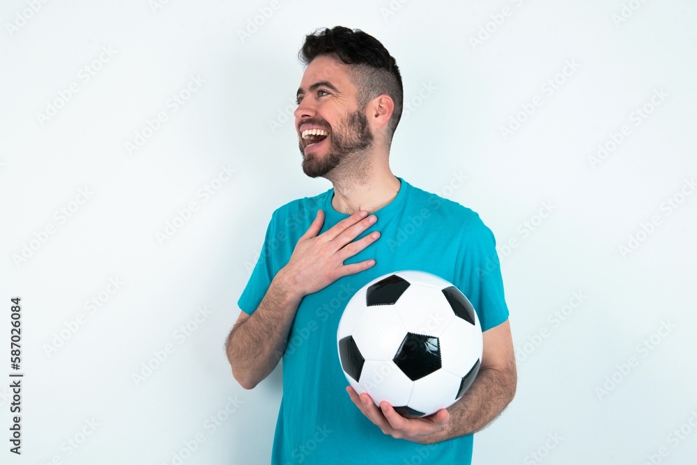 Joyful Young man holding a ball over white background expresses positive emotions recalls something funny keeps hand on chest and giggles happily. Joyful Young man holding a ball over white background expresses positive emotions recalls something funny keeps hand on chest and giggles happily.