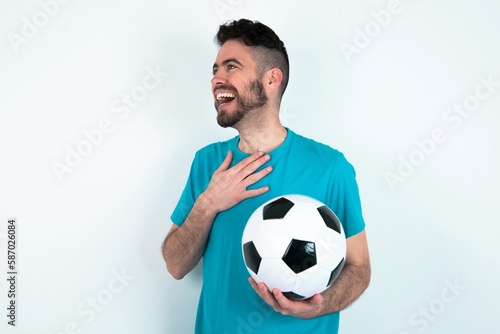 Joyful Young man holding a ball over white background expresses positive emotions recalls something funny keeps hand on chest and giggles happily.