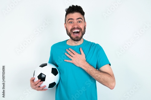 Young man holding a ball over white background smiles toothily cannot believe eyes expresses good emotions and surprisement