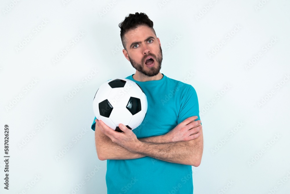 Shocked embarrassed Young man holding a ball over white background keeps mouth widely opened. Hears unbelievable novelty stares in stupor