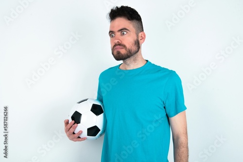 Young man holding a ball over white background stares aside with wondered expression has speechless expression. Embarrassed model looks in surprise