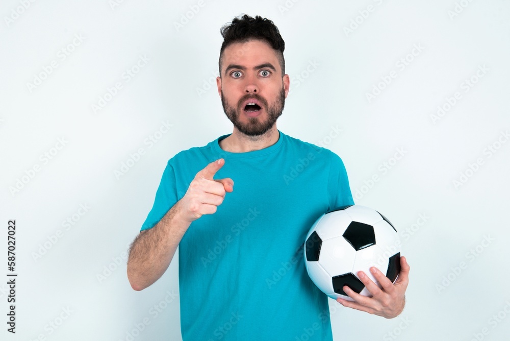 Naklejka premium Shocked Young man holding a ball over white background points at you with stunned expression