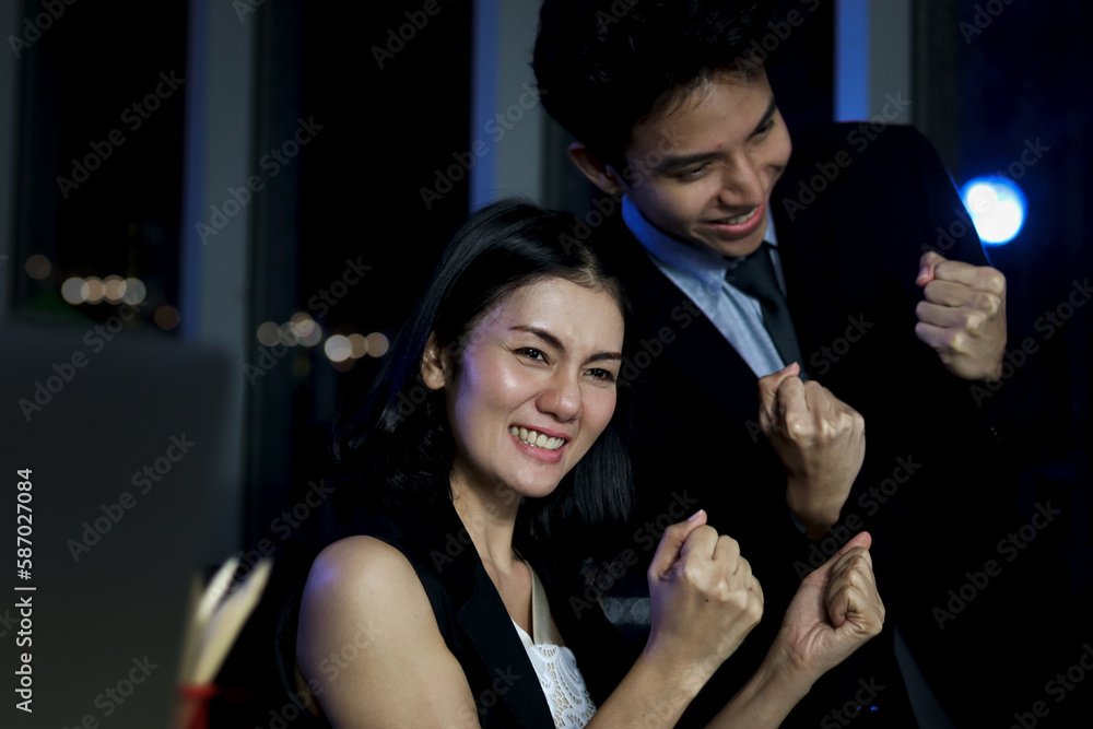 Cheerful happy Asian staff officer business man and woman reaching hands up to celebrate after finishing working-hard overtime together at night, night shift worker have overwork project overnight