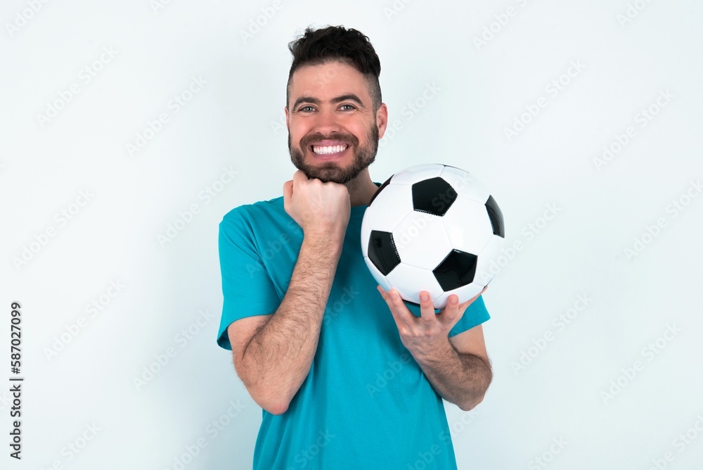 Satisfied Young man holding a ball over white background touches chin with both hands, smiles pleasantly, rejoices good day with lover