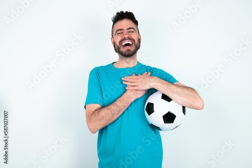 Young man holding a ball over white background expresses happiness, laughs pleasantly, keeps hands on heart