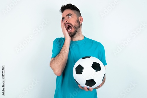 Young man holding a ball over white background keeps hands on cheeks has bored displeased expression. Stressed hopeless model