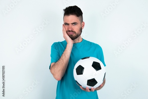 Sad lonely Young man holding a ball over white background touches cheek with hand bites lower lip and gazes with displeasure. Bad emotions