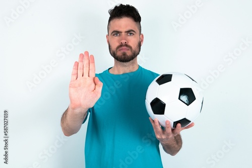 Young man holding a ball over white background shows stop sign prohibition symbol keeps palm forward to camera with strict expression