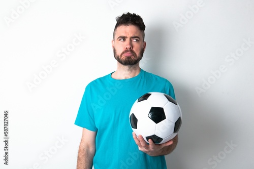 Dissatisfied Young man holding a ball over white background purses lips and has unhappy expression looks away stands offended. Depressed frustrated model.