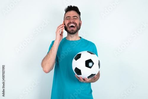 Funny Young man holding a ball over white background laughs happily, has phone conversation, being amused by friend, closes eyes.