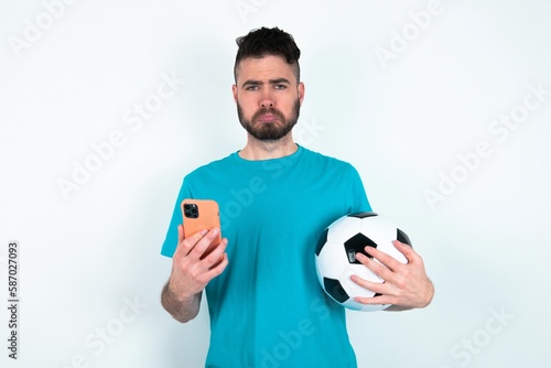 Upset dissatisfied Young man holding a ball over white background uses mobile software application and surfs information in internet, holds modern mobile hand