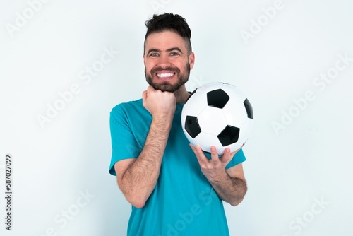 Satisfied Young man holding a ball over white background touches chin with both hands, smiles pleasantly, rejoices good day with lover