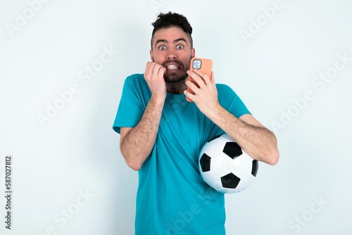 Afraid funny Young man holding a ball over white background holding telephone and bitting nails