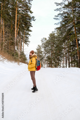 Young woman traveler with backpack and yellow coat walking on snow covered road in winter forest in frosty weather.