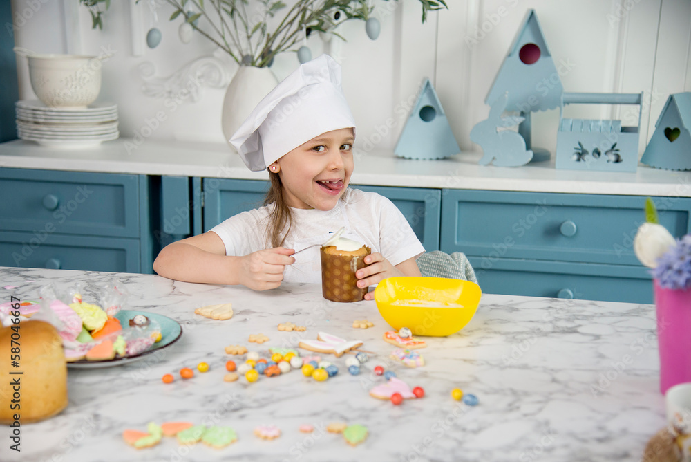 little cute funny smiling girl wearing a white chef's hat and uniform holding decorating easter cake in the blue and white kitchen with easter decoration