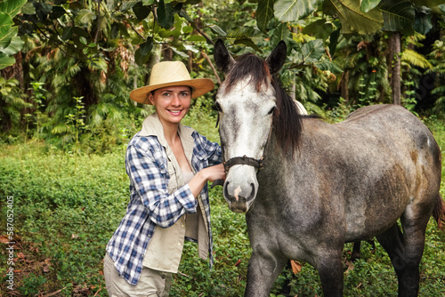 Young woman in shirt and straw hat posing next to gray horse, jungle trees background - horseriding, ranch at Isalo Park, Madagascar © Lubo Ivanko