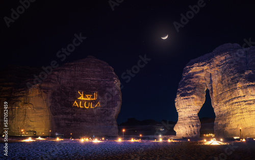 Jabal AlFil - Elephant Rock in evening, landscape illuminated, text Alula and Arabic translation projected to stone wall seats for people set up on the ground, crescent moon - composite image - above photo