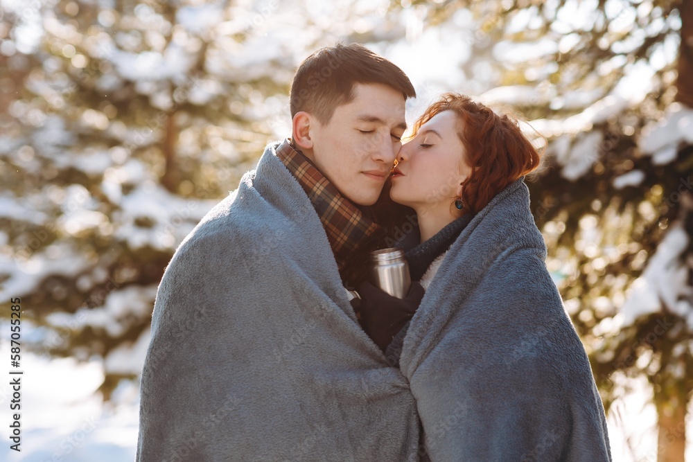 Fototapeta premium Close-up shot of a young couple in love in the middle of a snowy forest. A married couple is standing against the backdrop of snow-covered trees wrapped in a blanket. Young people have a date in