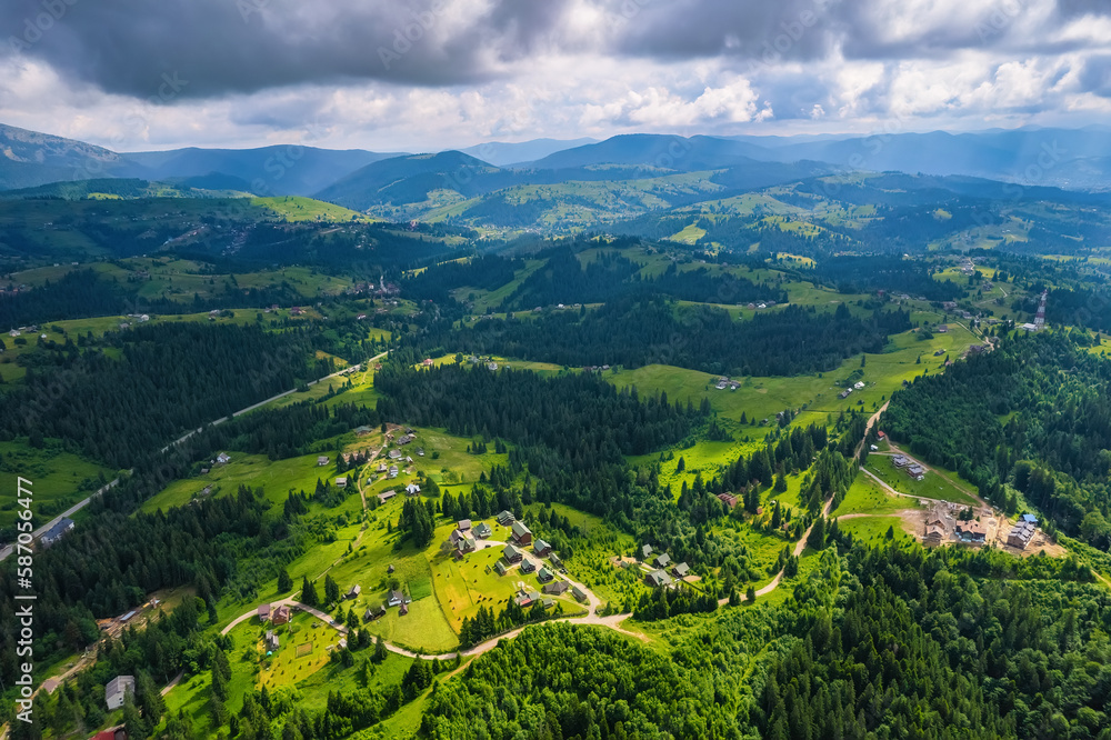 Scenic aerial view of the Carpathian mountains, village and blue sky with sun and clouds in morning light, summer rural landscape