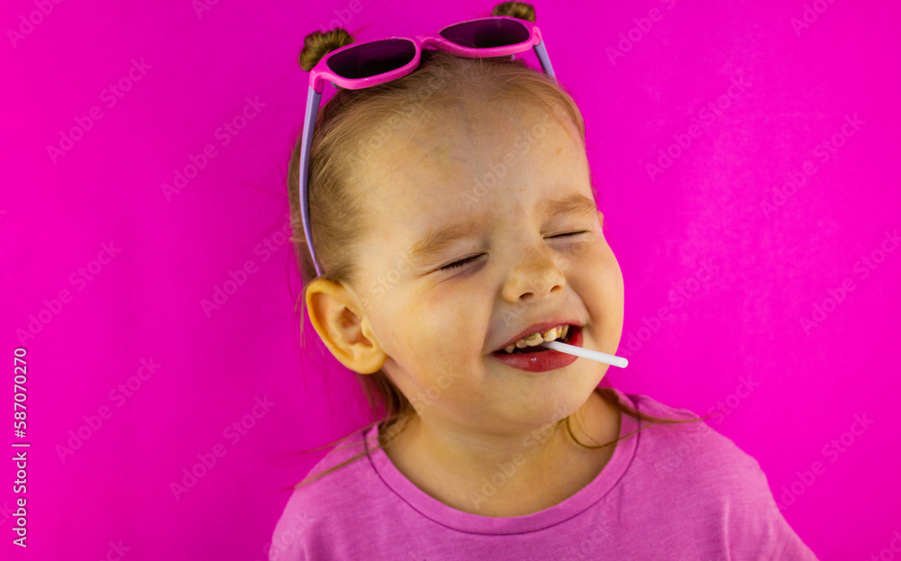 Beautiful cute baby girl screwed up her eyes with a lollipop in her mouth on a pink background. The child smiles happily in a candy. The concept of a cute baby screwed up his eyes with a lollipop. Beautiful cute baby girl screwed up her eyes with a lollipop in her mouth on a pink background. The child smiles happily in a candy. The concept of a cute baby screwed up his eyes with a lollipop.