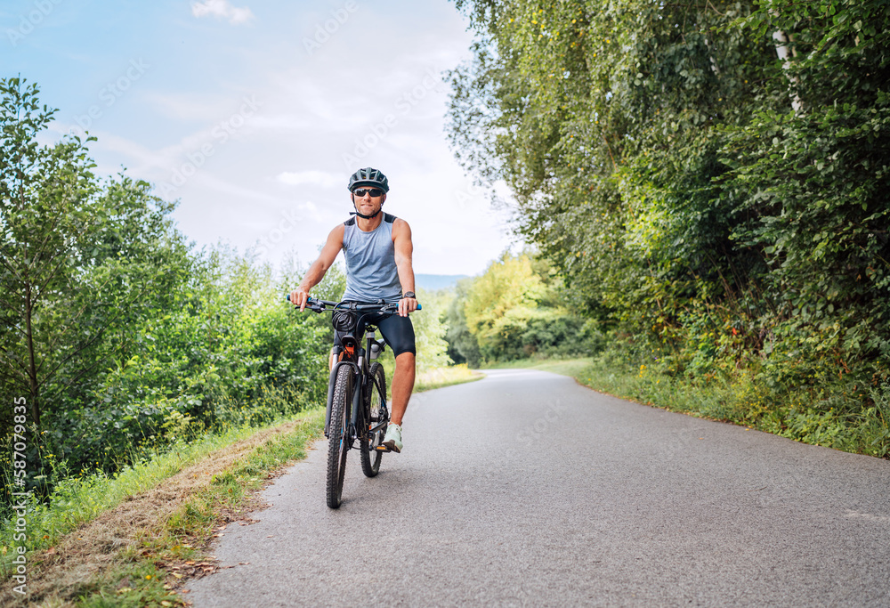 Portrait of a happy smiling man dressed in cycling clothes, helmet and sunglasses riding a bicycle on the asphalt out-of-town bicycle path. Active sporty people concept image.