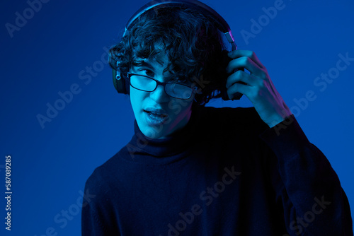 Teenage male with headphones listening to music and dancing and singing with glasses, hipster lifestyle, blue background, neon light, style and trends, mixed light, copy space © SHOTPRIME STUDIO