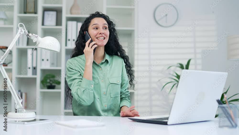Young attractive hispanic latin woman talking on the phone while sitting in modern office at workplace. Positive Smiling Curly Brunette Businesswoman Communicating Using Smartphone Indoor