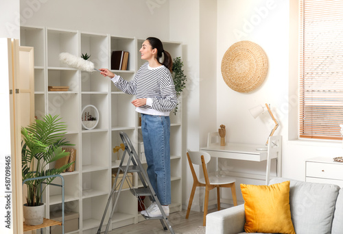 Foto Young woman on stepladder cleaning bookshelf with duster at home