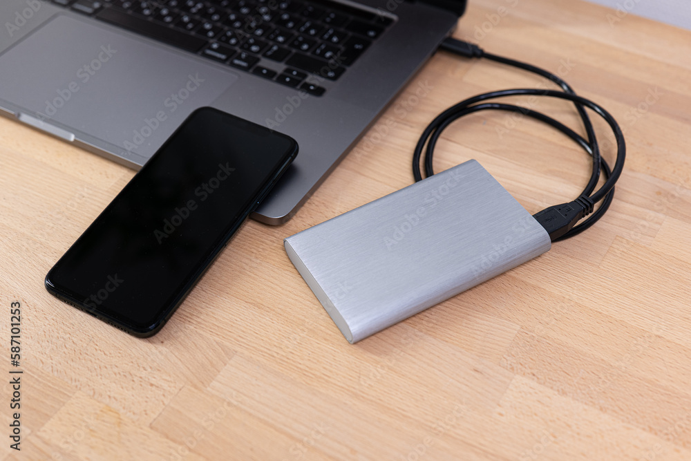 External hard drive with laptop computer and black phone. Portable hard drive connected to a laptop computer on a wooden table. Data transfer or backup between a laptop and a hard disk. External hard drive with laptop computer and black phone. Portable hard drive connected to a laptop computer on a wooden table. Data transfer or backup between a laptop and a hard disk.