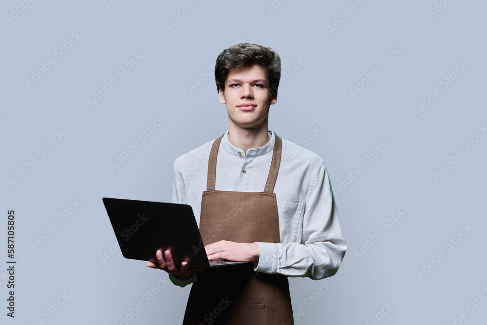 Young male in an apron with laptop, on gray studio background