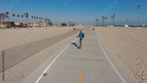 aerial footage of an African American man riding a OneWheel electric skateboard on a bike path with ocean water, brown sand, blue sky and kites flying at Belmont Shore Beach in Long Beach California