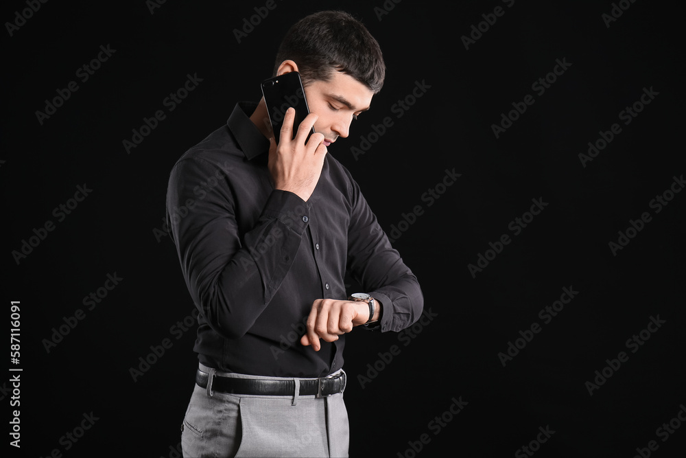 Business consultant talking by mobile phone and looking at wristwatch on dark background