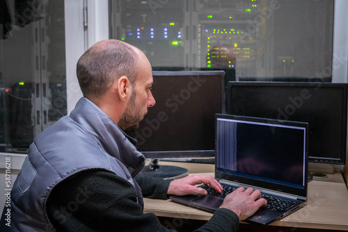 A man works on a laptop next to a server room. A programmer sits at a table in a data center.