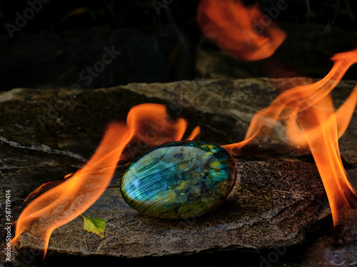 Ein rundlicher Labradorit liegt auf einer Schiefplatte. Der Edelstein schimmert in blau und grün und ist von Feuer umgeben. Der Hintergrund verschwindet in der Dunkelheit. photo