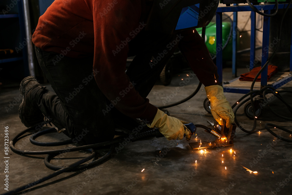A hardworking mechanic in a welding mask and oil-stained clothing is welding parts in an auto repair shop or car workshop. Blue flashes of light and bright orange sparks are flying in all directions. A hardworking mechanic in a welding mask and oil-stained clothing is welding parts in an auto repair shop or car workshop. Blue flashes of light and bright orange sparks are flying in all directions.