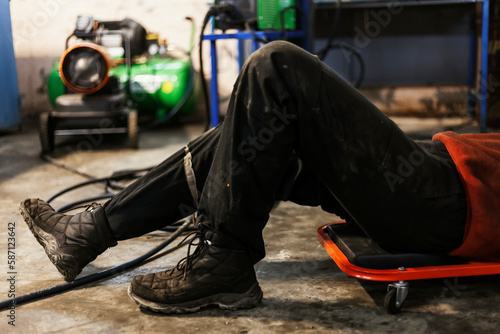 A hardworking mechanic with oil-stained clothing on a mechanics creeper is inspecting a bottom of the car. legs close-up.