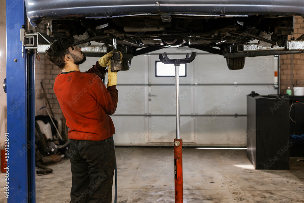 A hardworking mechanic with oil-stained clothing is working with a pneumatic impact wrench underneath the bottom of a car that has been lifted on an electric lift.