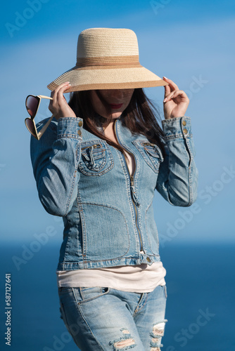 Portrait of hipster woman in denim jacket and jeans hiding with straw hat over his face. Adult female posing on background of blue cloudless sky and ocean on sunny day. Summer travel concept