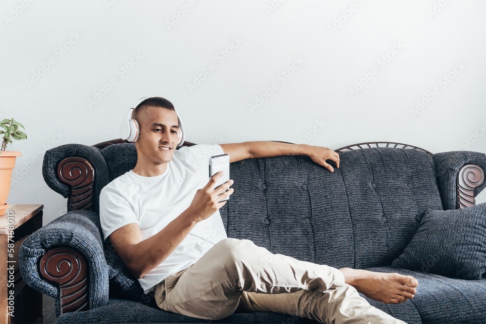 happy young hispanic man listen music with headphones lying down on sofa.