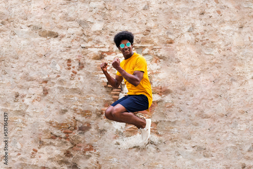 Happy young black man jumping in the air. Rustic brick and stone wall background.