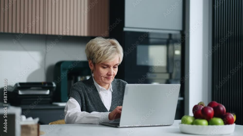 Charming Senior Woman Typing Text On Keyboard Of Laptop In Home Kitchen, Portrait Indoors Charming Senior Woman Typing Text On Keyboard Of Laptop In Home Kitchen, Portrait Indoors