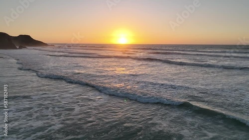 Beautiful droneshot of portugese beach with powerful sunset on the horizon. Waves rolling into the sandy beach. Surrounded by large rocky cliffs.