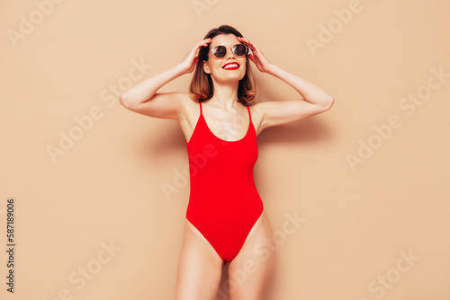 Young beautiful sexy woman. Smiling carefree model wearing red lingerie. Hot tanned female posing near beige wall in studio in summer swimwear bathing suit. In sunglasses. slim and fit