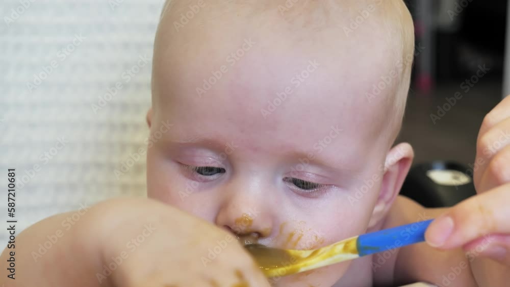 Mom feeding little boy with broccoli puree. Child at the age of six months eats broccoli while sitting on a baby chair. Slow motion shot. Mom feeding little boy with broccoli puree. Child at the age of six months eats broccoli while sitting on a baby chair. Slow motion shot.