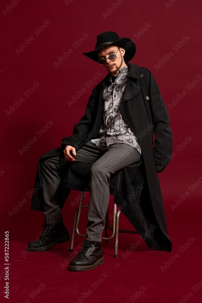 Young man sitting in a chair, wearing a hat, overcoat, and sunglasses on a red background Young man sitting in a chair, wearing a hat, overcoat, and sunglasses on a red background
