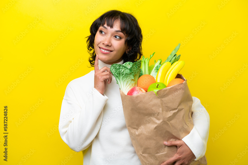 Young Argentinian woman holding a grocery shopping bag isolated on yellow background thinking an idea while looking up