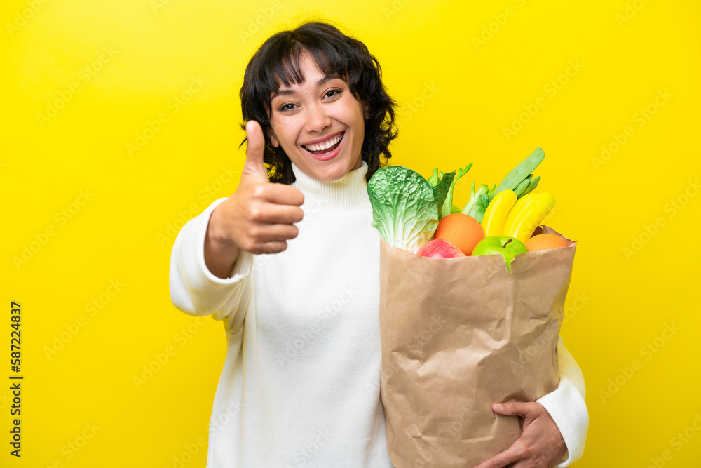 Young Argentinian woman holding a grocery shopping bag isolated on yellow background with thumbs up because something good has happened Young Argentinian woman holding a grocery shopping bag isolated on yellow background with thumbs up because something good has happened