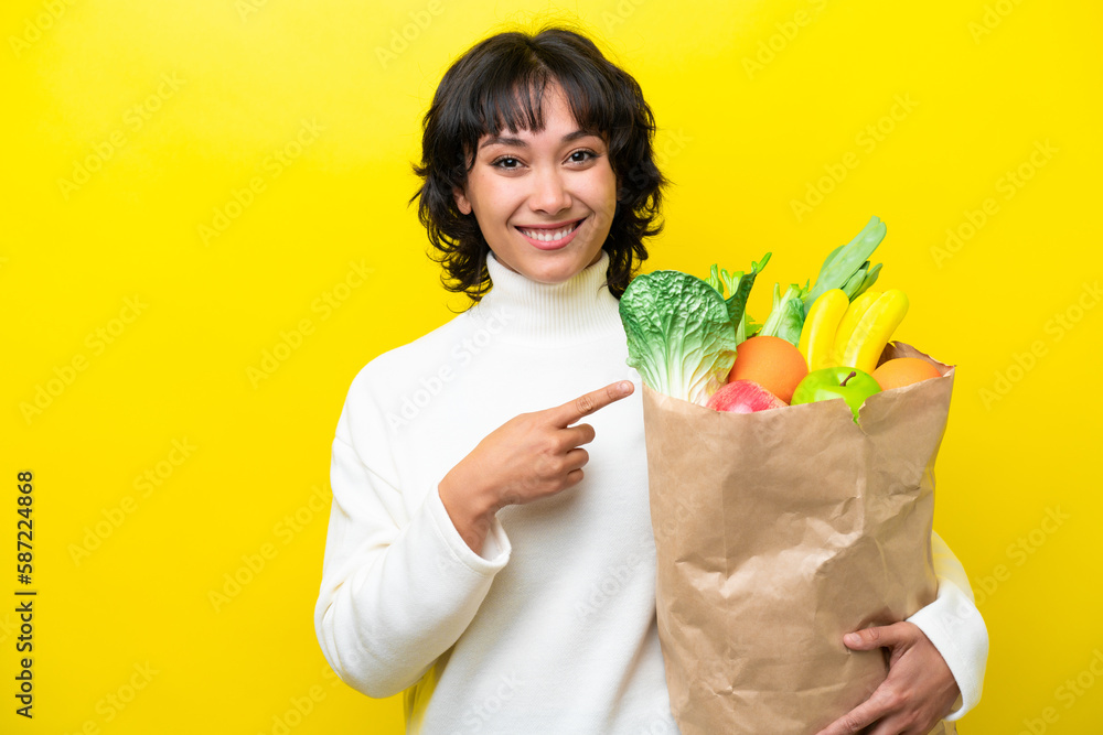 Young Argentinian woman holding a grocery shopping bag isolated on yellow background pointing to the side to present a product Young Argentinian woman holding a grocery shopping bag isolated on yellow background pointing to the side to present a product