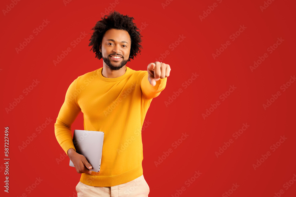 Emotional man pointing you and holding laptop while looking at camera with happy smile. Indoor studio shot isolated on red background Emotional man pointing you and holding laptop while looking at camera with happy smile. Indoor studio shot isolated on red background