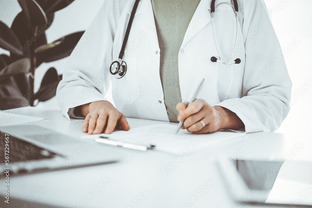 Unknown doctor woman sitting and writing notes at the desk in clinic or hospital office, close up. Medicine concept. Unknown doctor woman sitting and writing notes at the desk in clinic or hospital office, close up. Medicine concept.